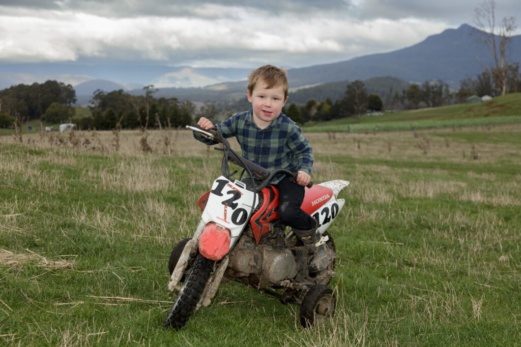Young boy smiling on a muddy Honda mini motorbike in a grassy field in rural Australia